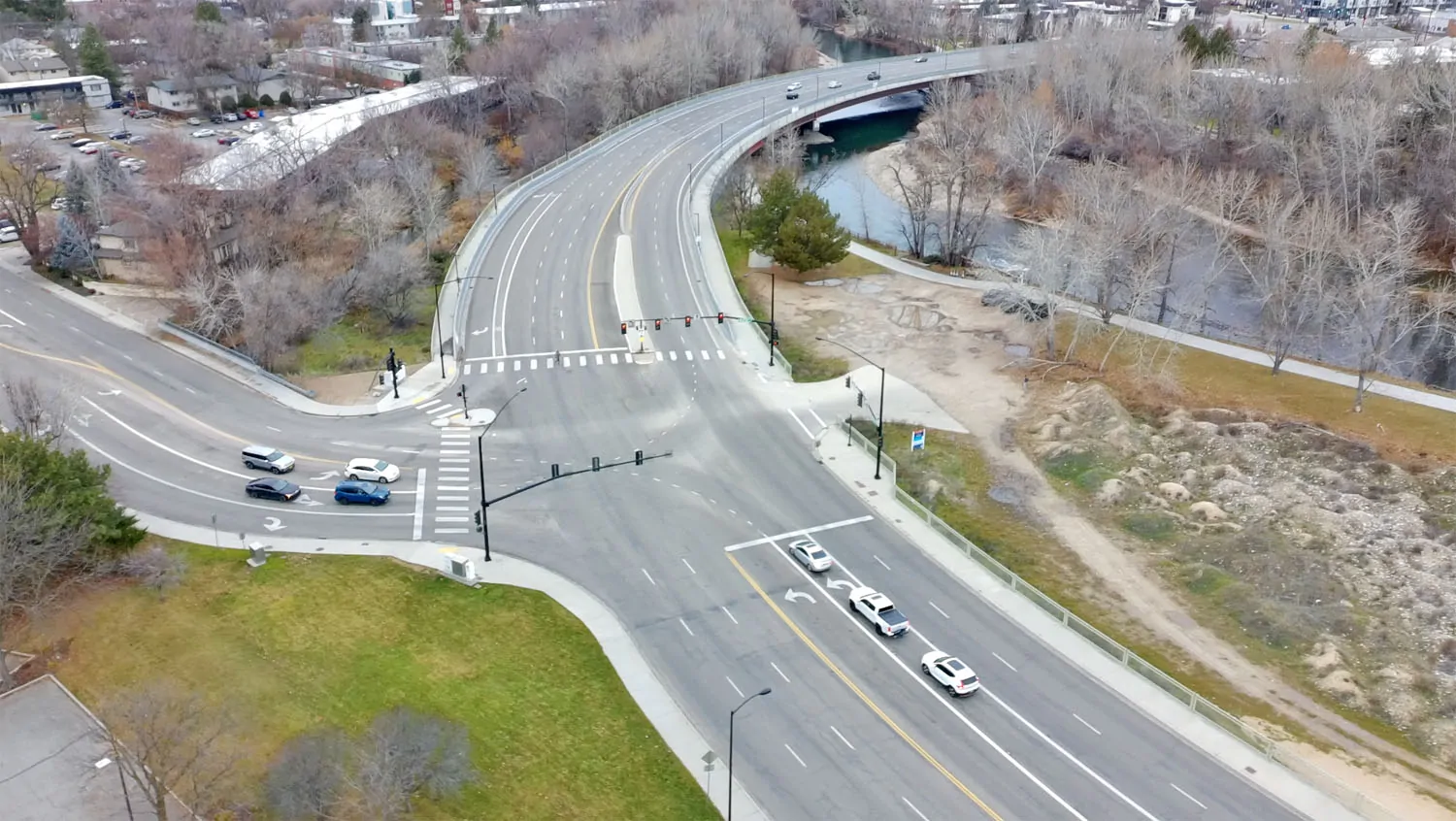 Parkcenter Before An aerial photograph of the Parkcenter bridge in Boise, Idaho, showing a multi-lane asphalt roadway and heavy concrete infrastructure crossing the Boise River, surrounded by utilitarian traffic lights and winter-toned vegetation.