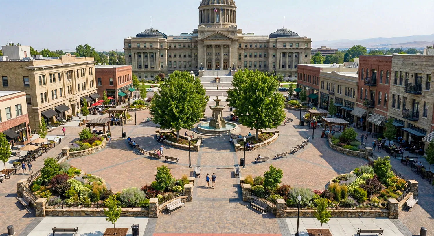 Pedestrian-oriented Capitol Square with trees, fountain, seating, and people gathered in a landscaped civic plaza in front of the Idaho State Capitol.