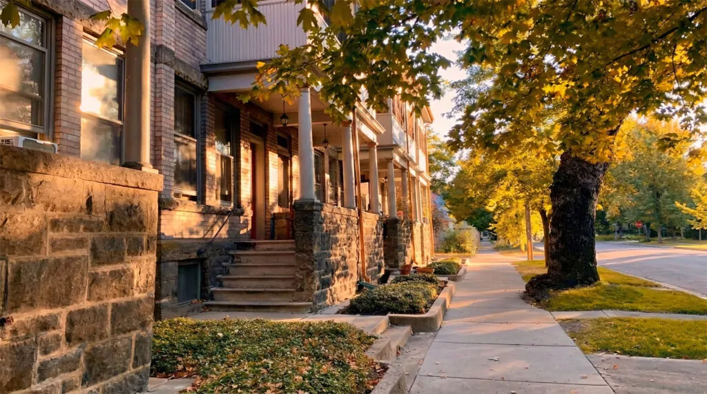 Historic brick and stone multifamily building in Boise's North End at golden hour. The wide, clean sidewalk is lined with mature trees and lush green groundcover, leading past classic columned porches under a warm dusk light.