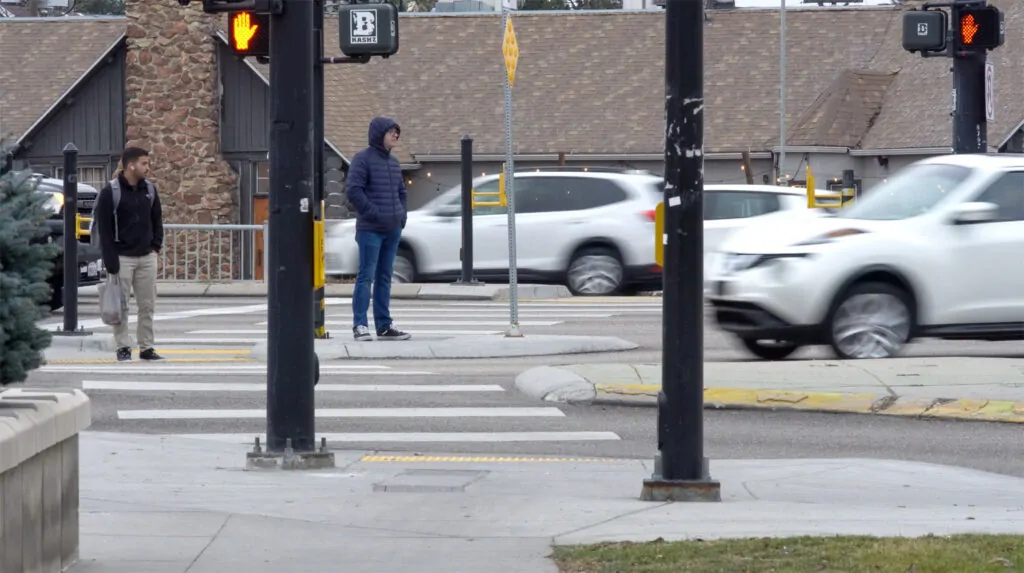 Pedestrians wait at a Boise crosswalk as traffic moves through a busy intersection, illustrating the tension between human movement and civic coordination.