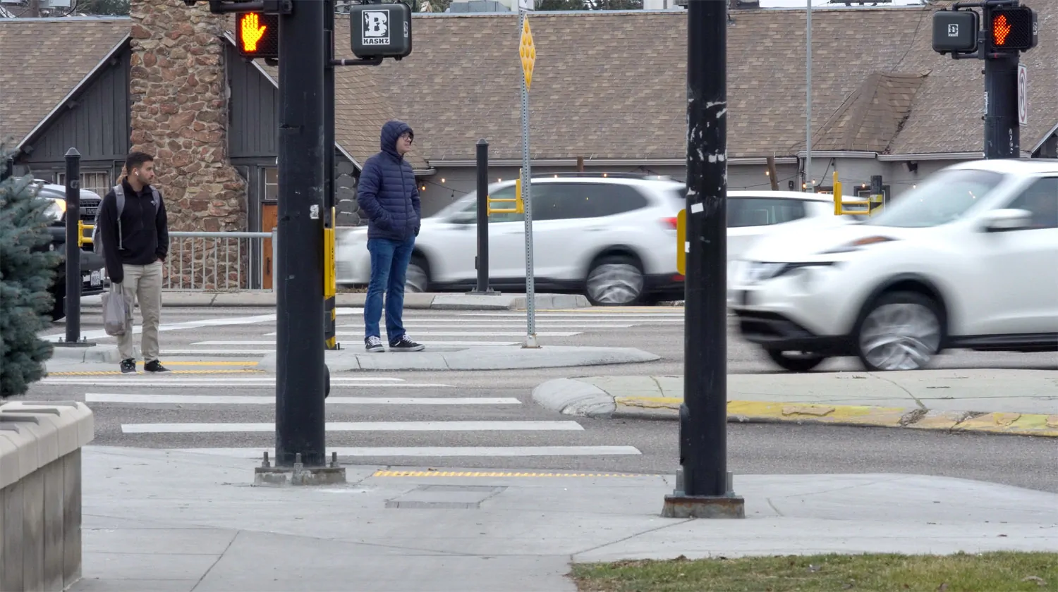 Pedestrians wait at a Boise crosswalk as traffic moves through a busy intersection, illustrating the tension between human movement and civic coordination.