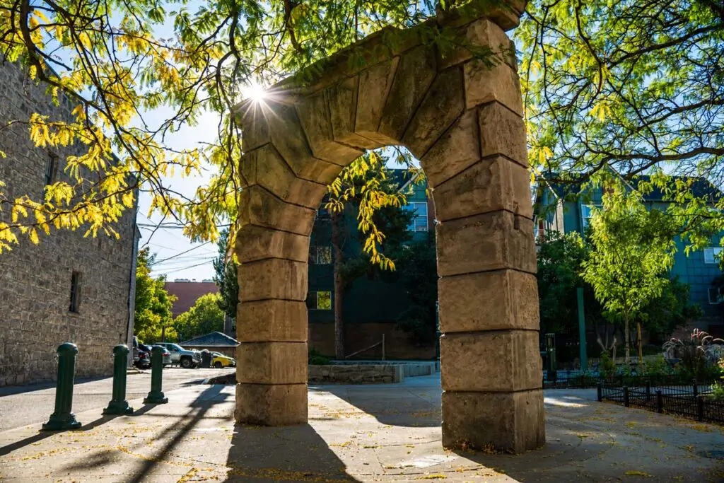 Sunlit photograph of a weathered stone archway in downtown Boise, framed by leafy tree branches and long shadows, with a bright starburst of light filtering through the canopy and nearby buildings and sidewalk visible in the background.