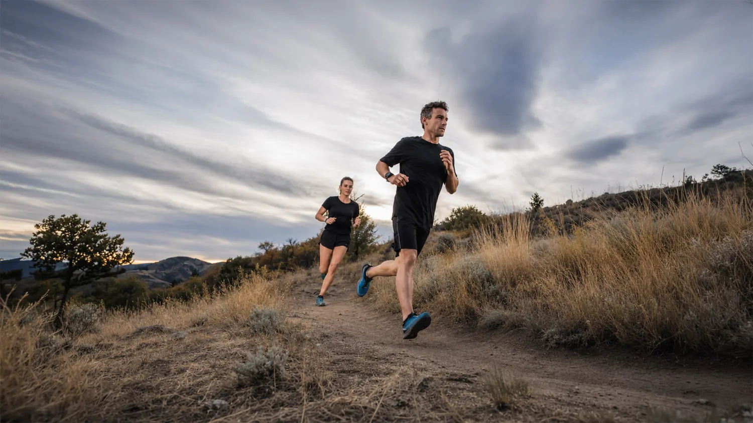 Two runners move along a dry foothills trail under a wide sky, suggesting effort, place, and the ritual of moving through landscape.