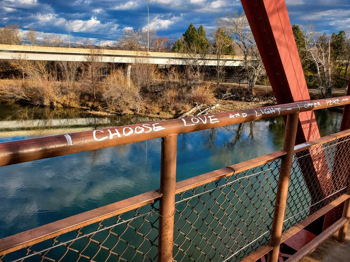 White graffiti on a metal bridge railing reads “I choose love and light,” with the Boise River and trees in the background.