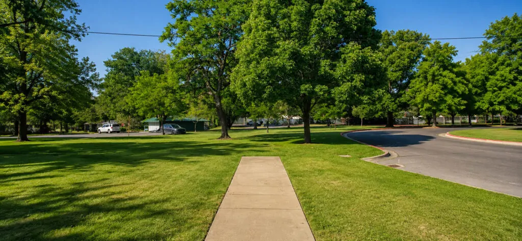 Wide view of a sidewalk ending abruptly in grass near Julia Davis Park in Boise, with trees, a curved road, and the park beyond.