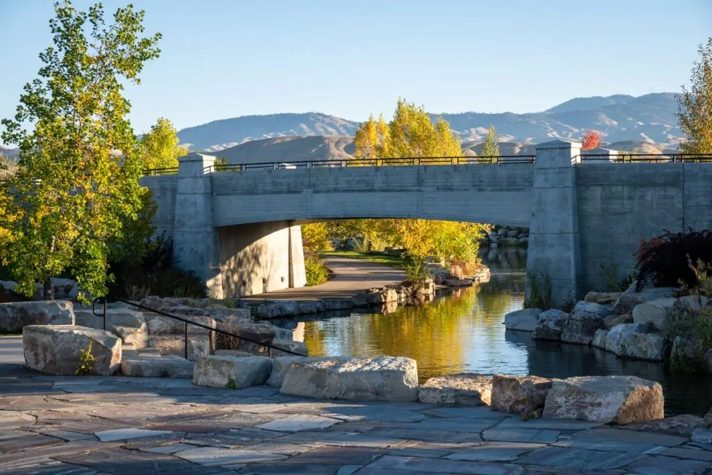 A pedestrian path passes beneath a concrete bridge beside a landscaped waterway, with trees and the Boise foothills in the distance.
