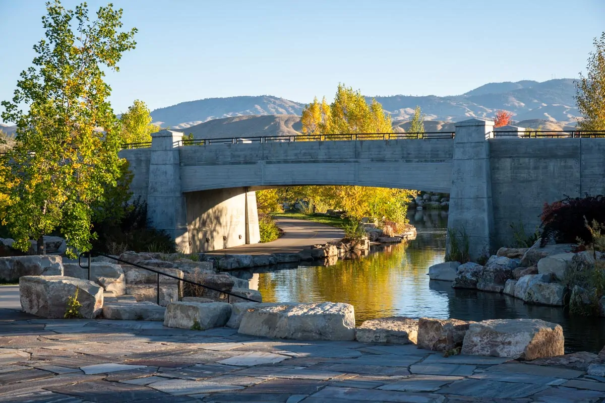 A pedestrian path passes beneath a concrete bridge beside a landscaped waterway, with trees and the Boise foothills in the distance.