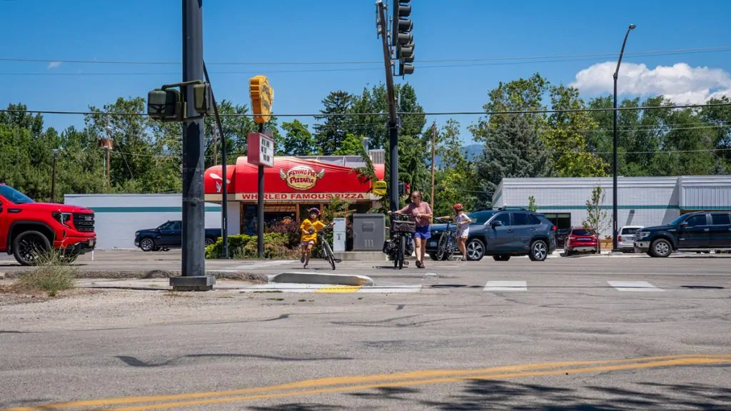 People with bicycles waiting at a crosswalk along a wide Boise street with cars and commercial buildings.